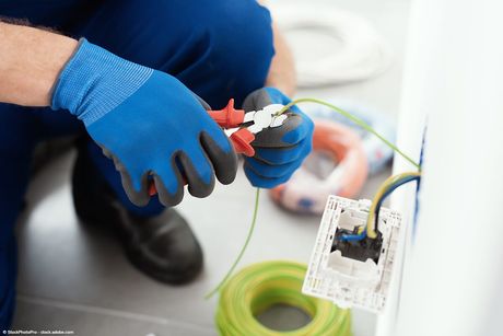 Ein Elektriker mit blauen Handschuhen arbeitet mit einer Zange an elektrischen Leitungen in der Nähe einer Steckdose.