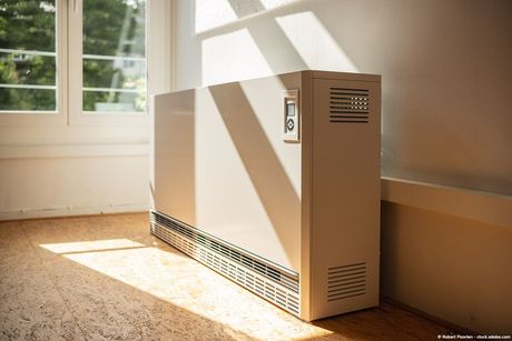 Ein beigefarbener Elektroheizkörper steht in einem Zimmer neben einer Wand. Sonnenlicht strömt durch ein Fenster.