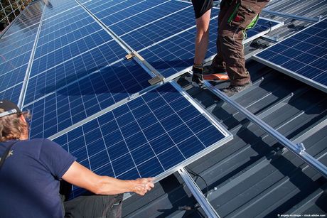 Arbeiter installieren an einem sonnigen Tag Solarpaneele auf einem grauen Metalldach.