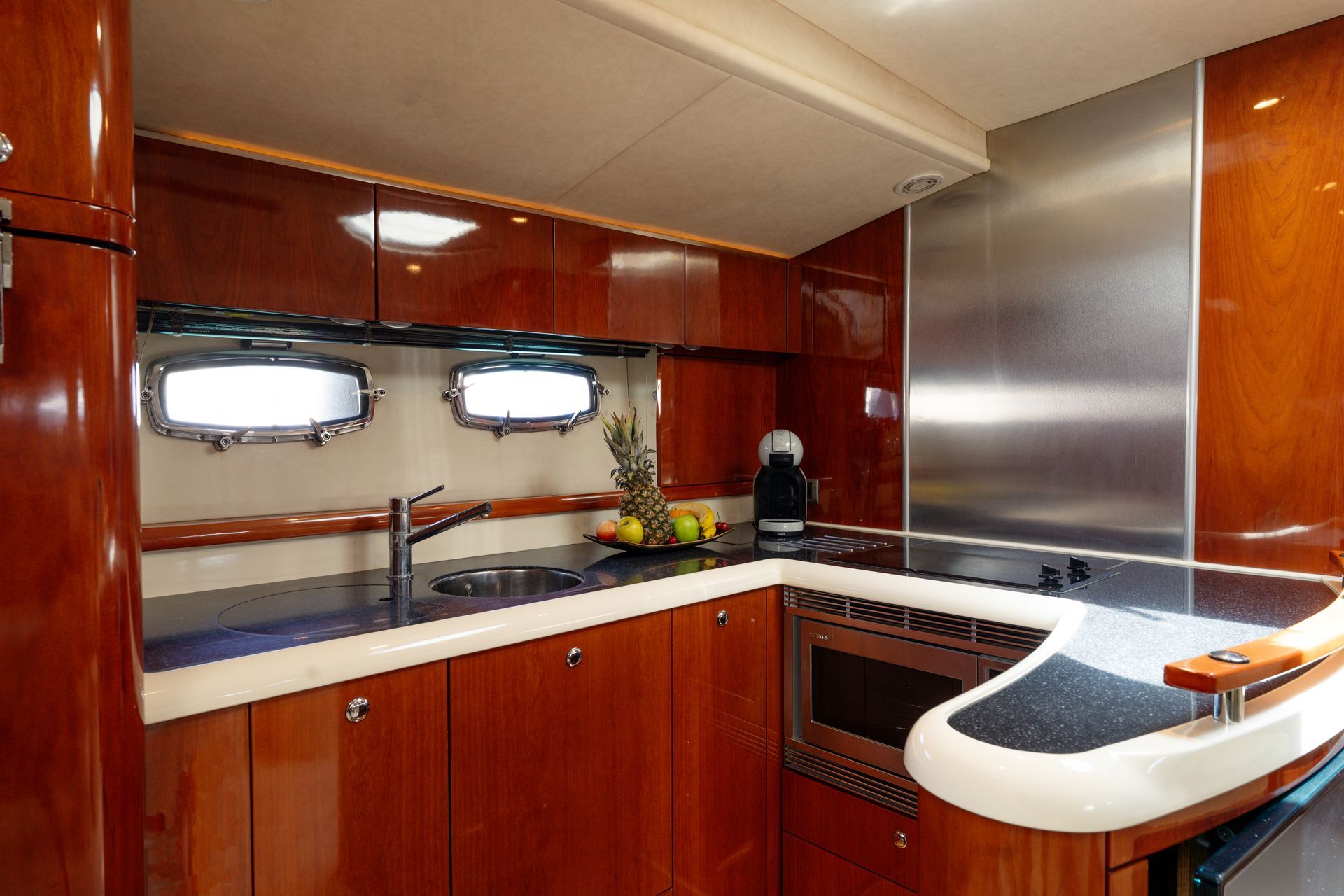 Compact yacht galley with wood cabinets, white countertop, stove, sink, and porthole windows.