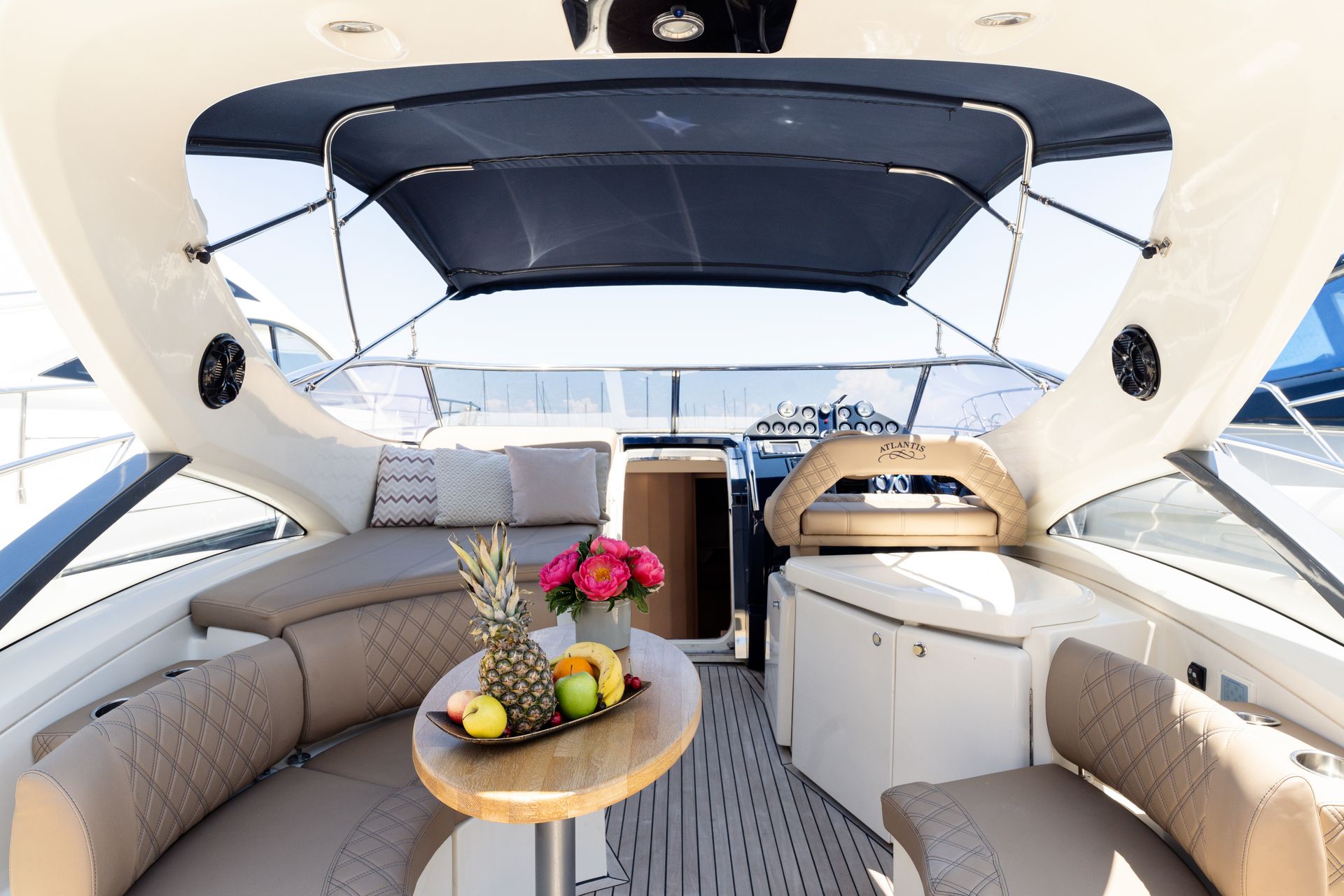 Luxury yacht cockpit with beige seating, round table with flowers and fruit, and a shaded canopy view.