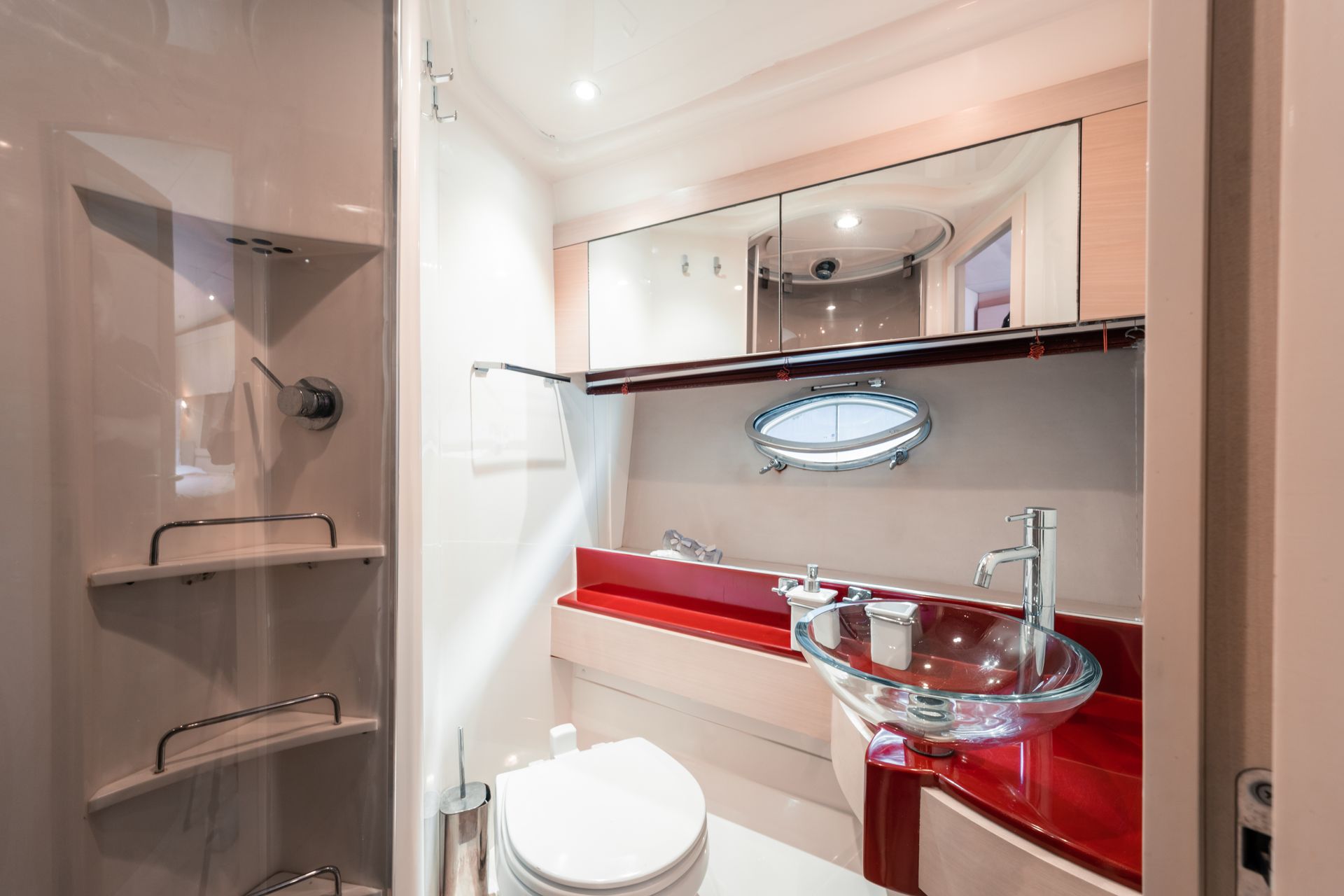 Small bathroom with toilet, glass sink, mirror, red counter, and shower shelf on the left