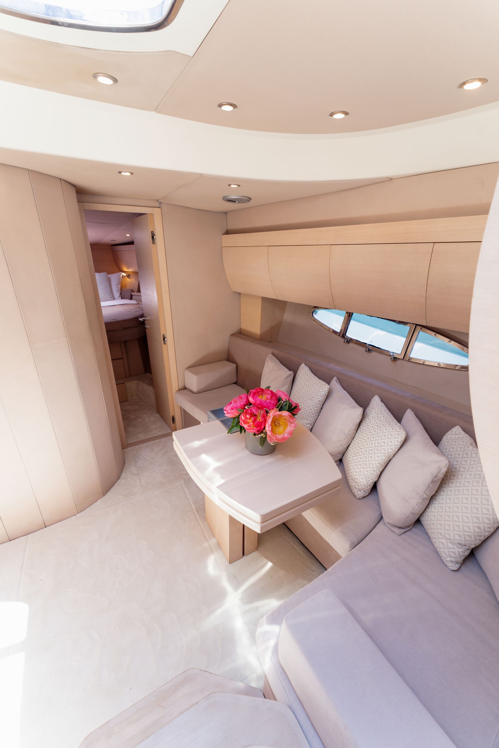 Compact yacht lounge with beige seating, a small table, flowers, and a view through side windows.