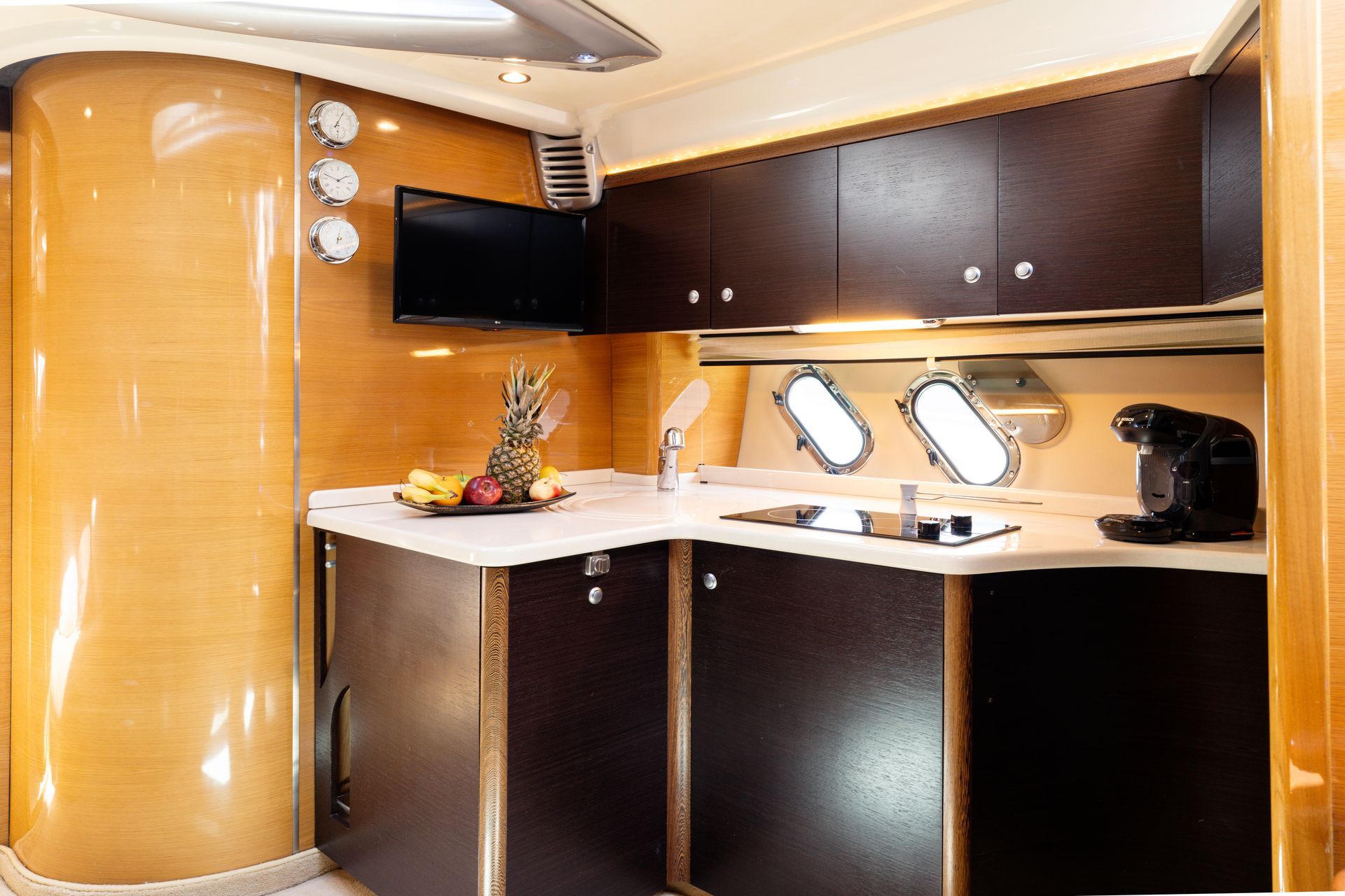 Modern yacht galley with wood paneling, white countertop, cabinets, TV, and porthole windows