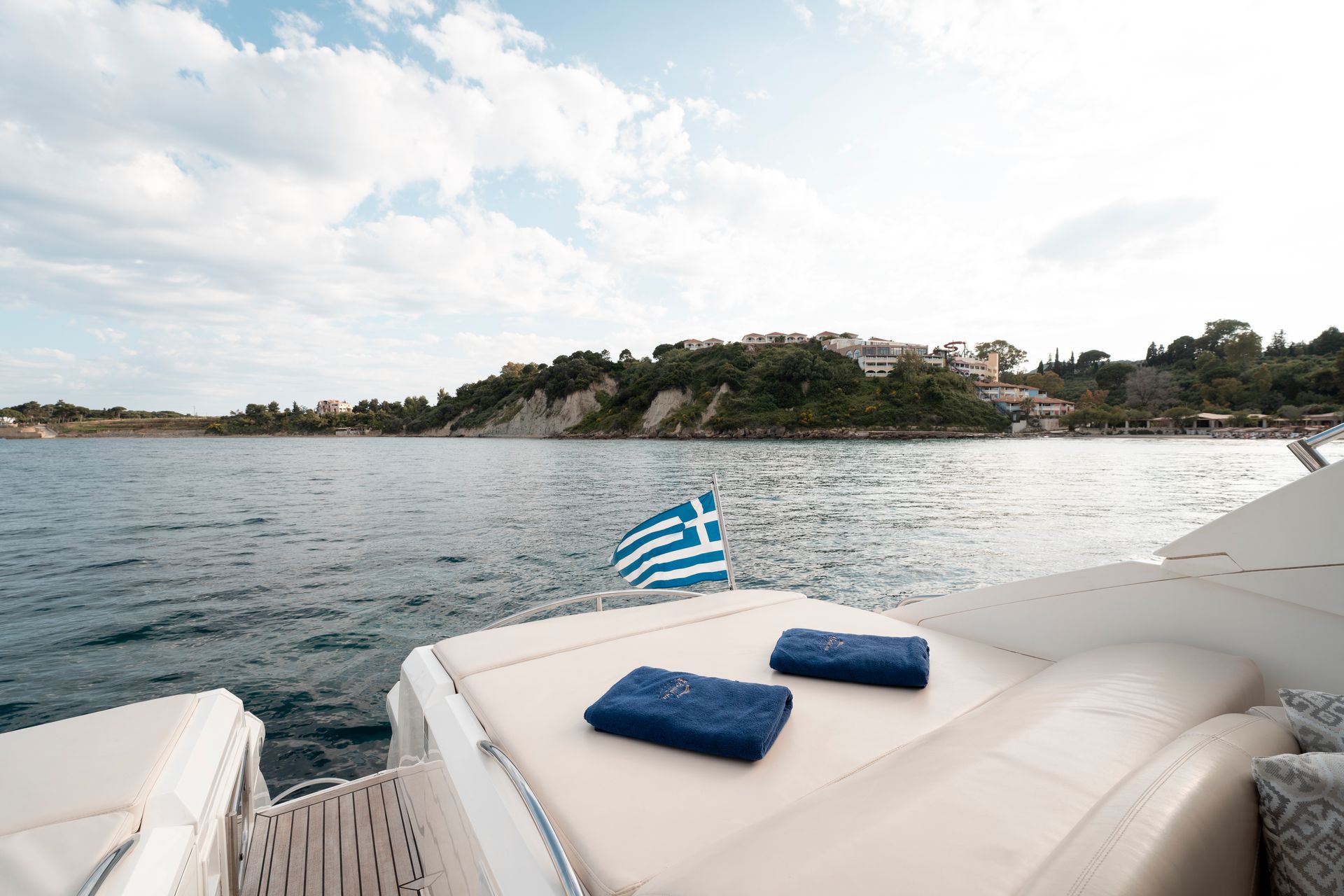 Yacht deck with blue towels and Greek flag, overlooking a calm sea and rocky coastline