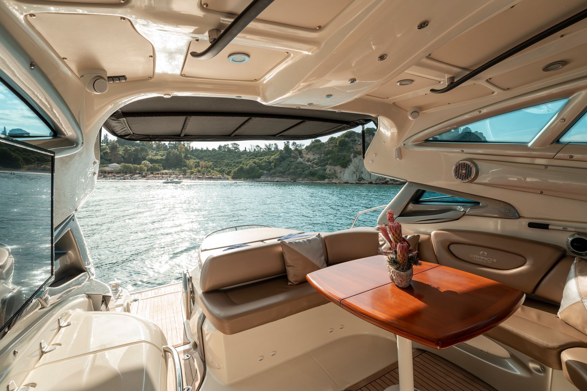 Yacht cabin with tan seats and wood table, open stern view to sunlit water and shoreline.