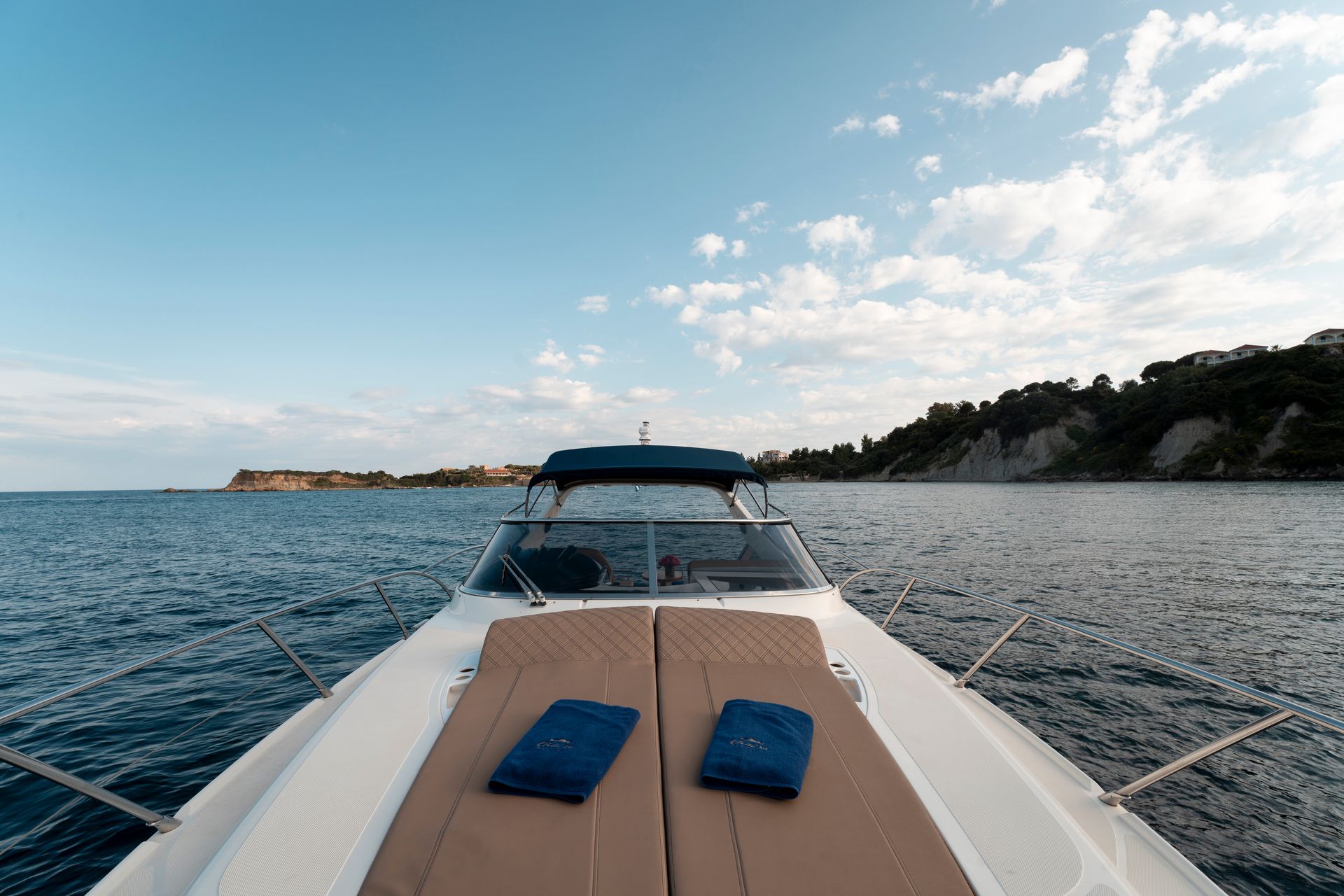 View from a boat’s bow on open water, with blue cushions and a rocky shoreline under a bright sky.