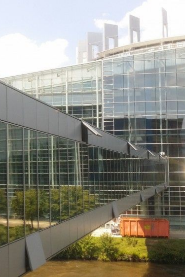 Passerelle vitrée surplombant un immeuble de bureaux moderne, avec une benne à ordures rouge en dessous.