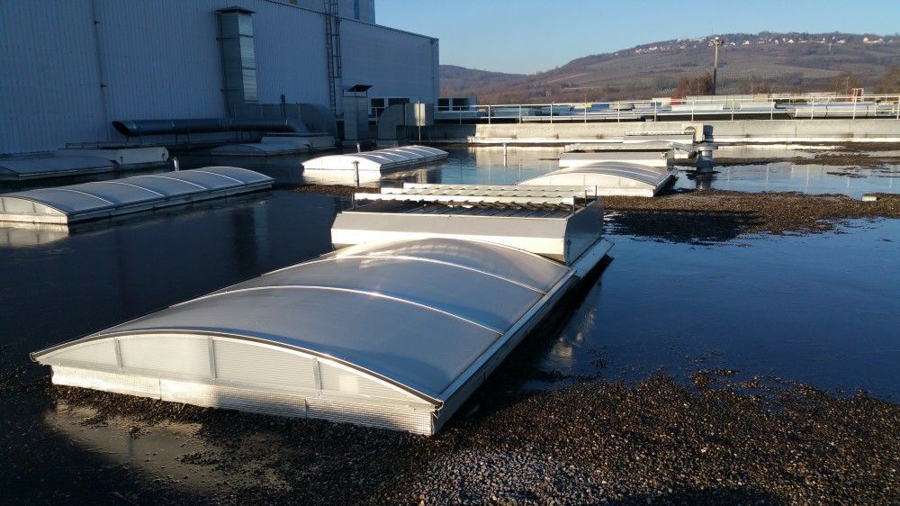 Des puits de lumière blancs sur le toit d'un bâtiment, au bord d'une étendue d'eau calme sous un ciel bleu limpide.