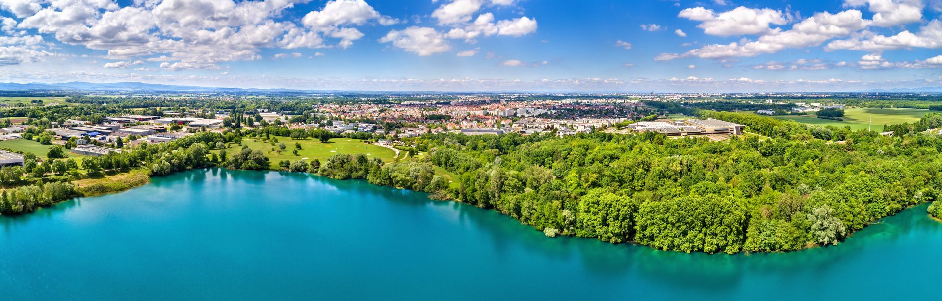 Vue aérienne panoramique d'un lac turquoise bordé d'une forêt verdoyante et d'une silhouette urbaine sous un ciel bleu.
