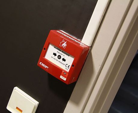 Boîtier d'alarme incendie rouge fixé au mur sur une colonne beige, avec un dispositif blanc en dessous.