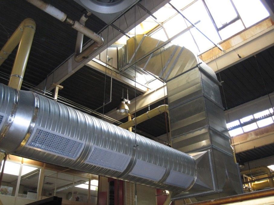 De larges conduits de ventilation argentés traversent un plafond industriel percé de puits de lumière et de poutres en acier.