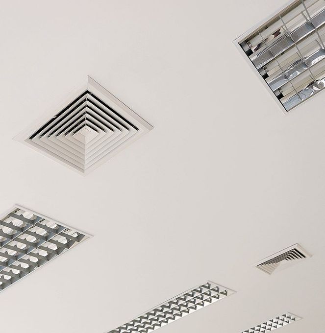 Plafond blanc avec plusieurs grilles de ventilation rectangulaires et panneaux lumineux fluorescents