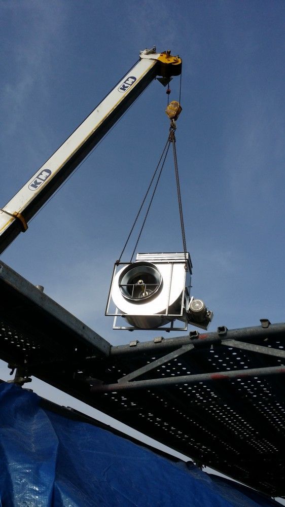 Une grue soulève une unité de climatisation extérieure à l'aide de chaînes, sur fond de ciel bleu.