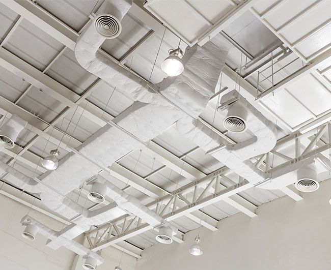 Plafond industriel blanc avec conduits, grilles d'aération, poutres et luminaires suspendus apparents.