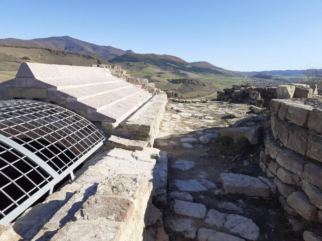 Un muro de piedra con una valla metálica que lo rodea y montañas al fondo.