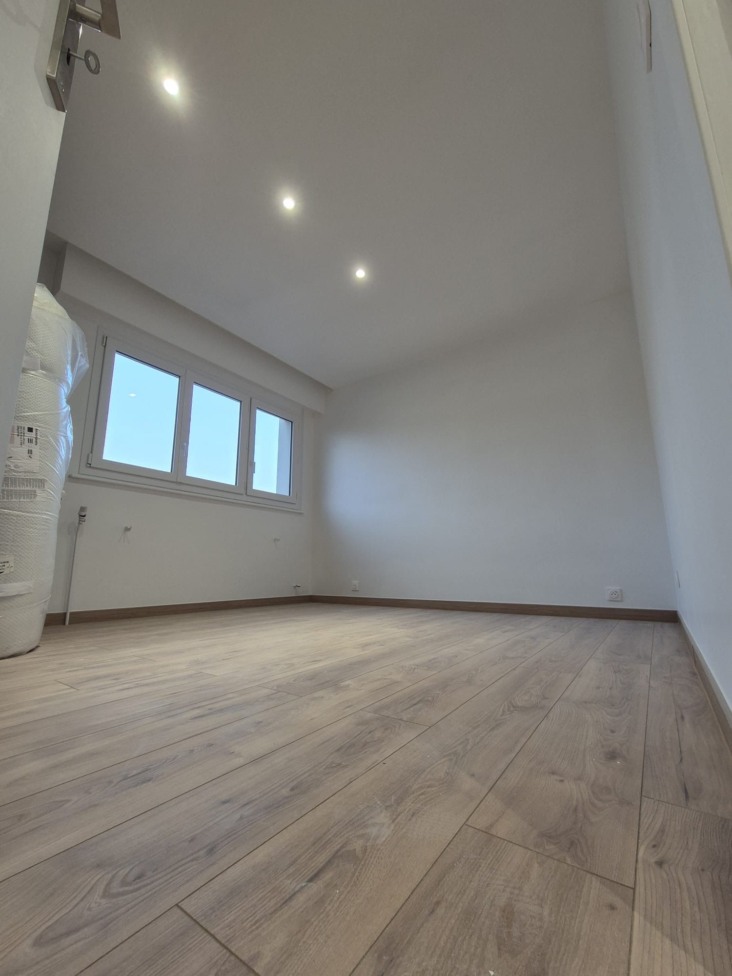 Pièce vide avec sol imitation bois, murs blancs, trois spots encastrés au plafond et une fenêtre.