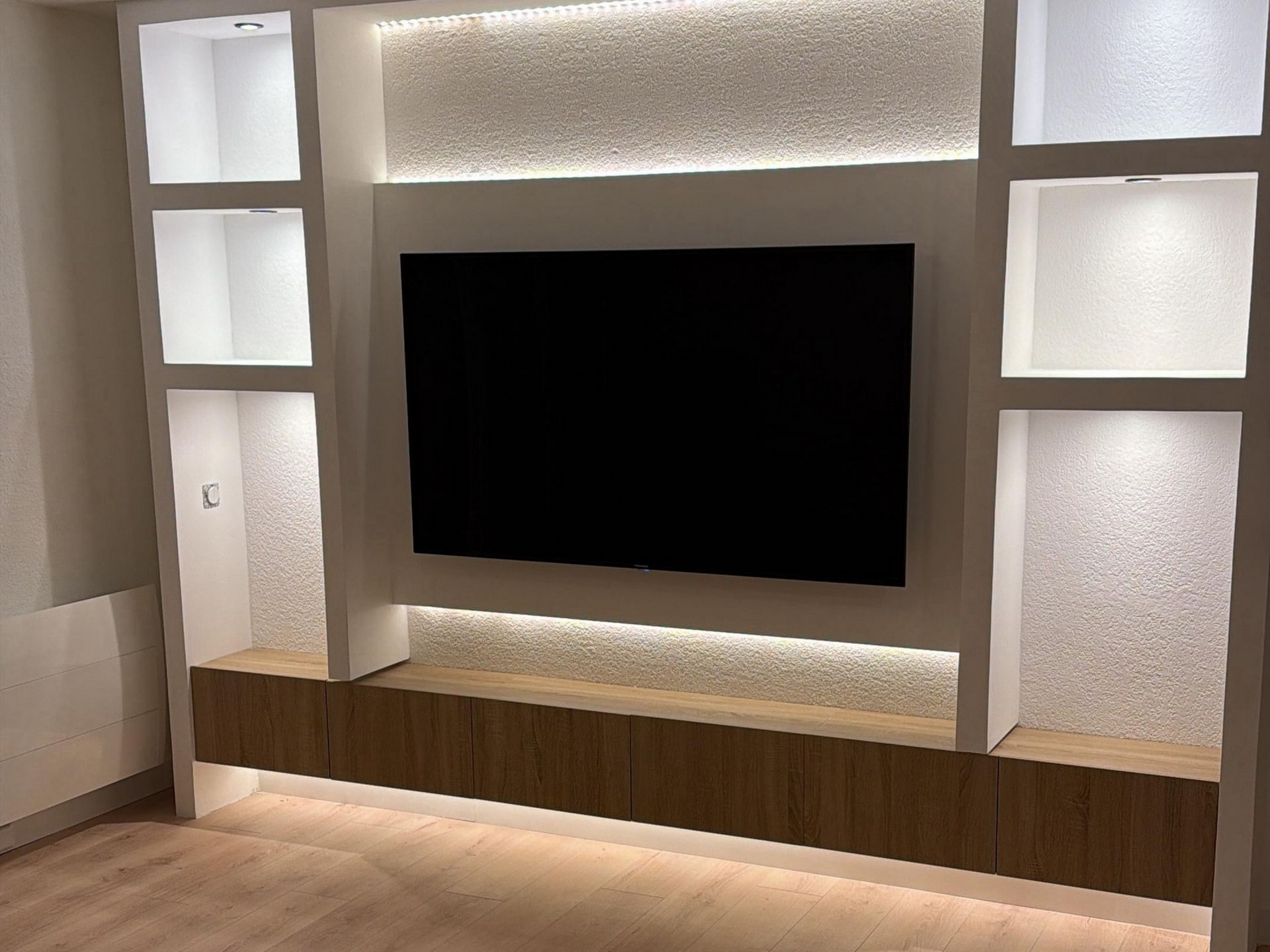 Meuble TV intégré avec télévision murale, étagères et placards en bois. Blanc avec accents texturés et éclairage LED.