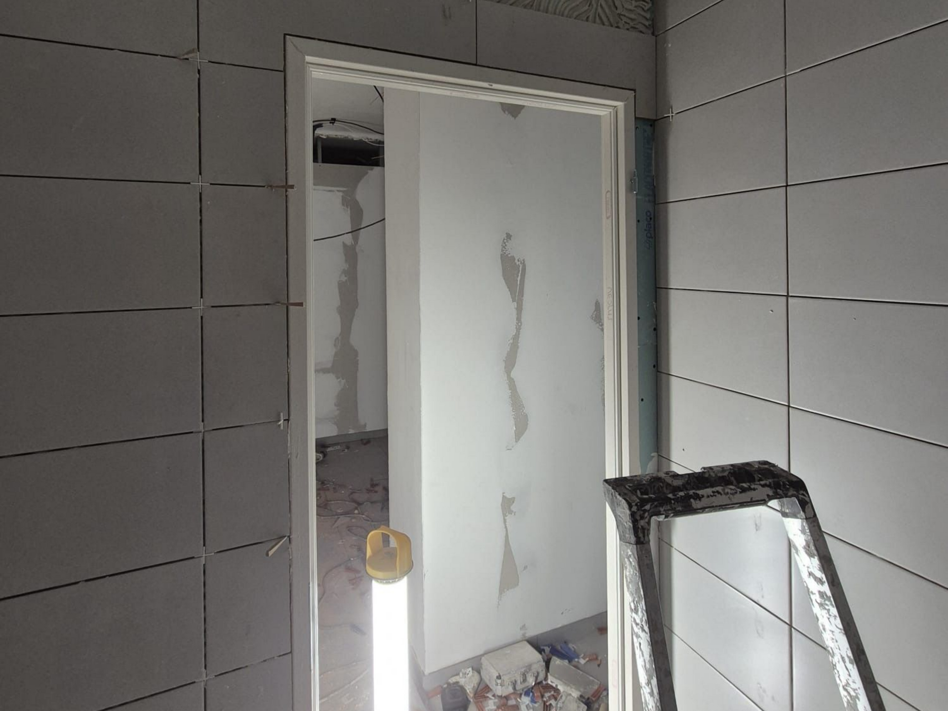 Salle de bains en construction, carrelée de blanc, avec porte ouverte. Une échelle est appuyée contre l'encadrement de la porte.