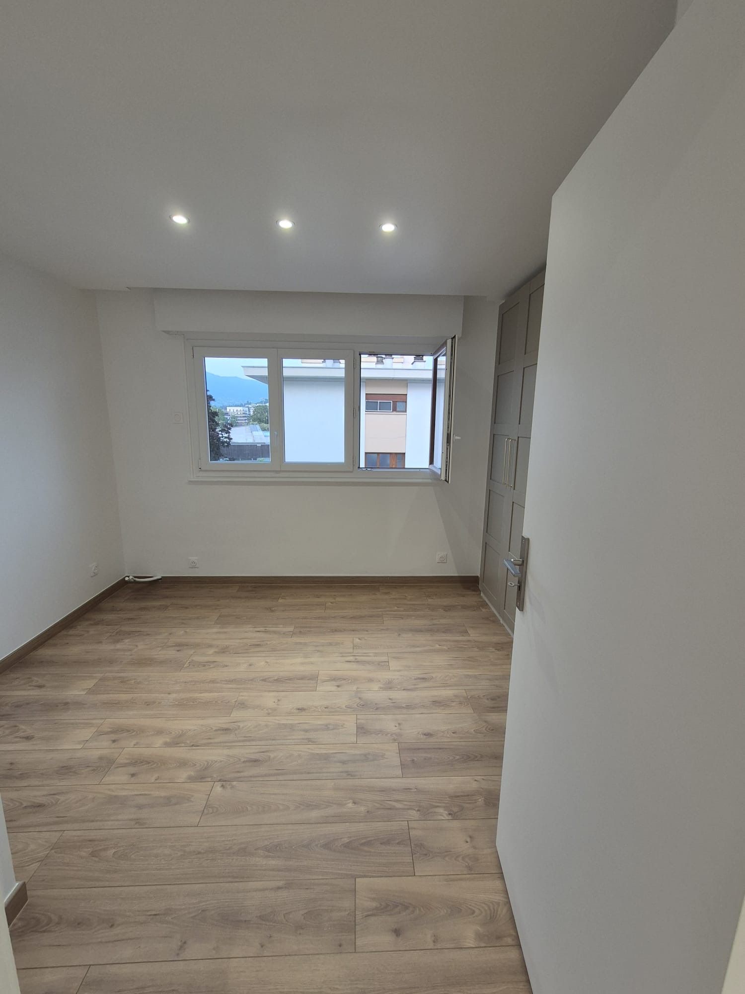 Pièce vide avec sol imitation bois, fenêtre et éclairage encastré. Une porte est ouverte au premier plan.