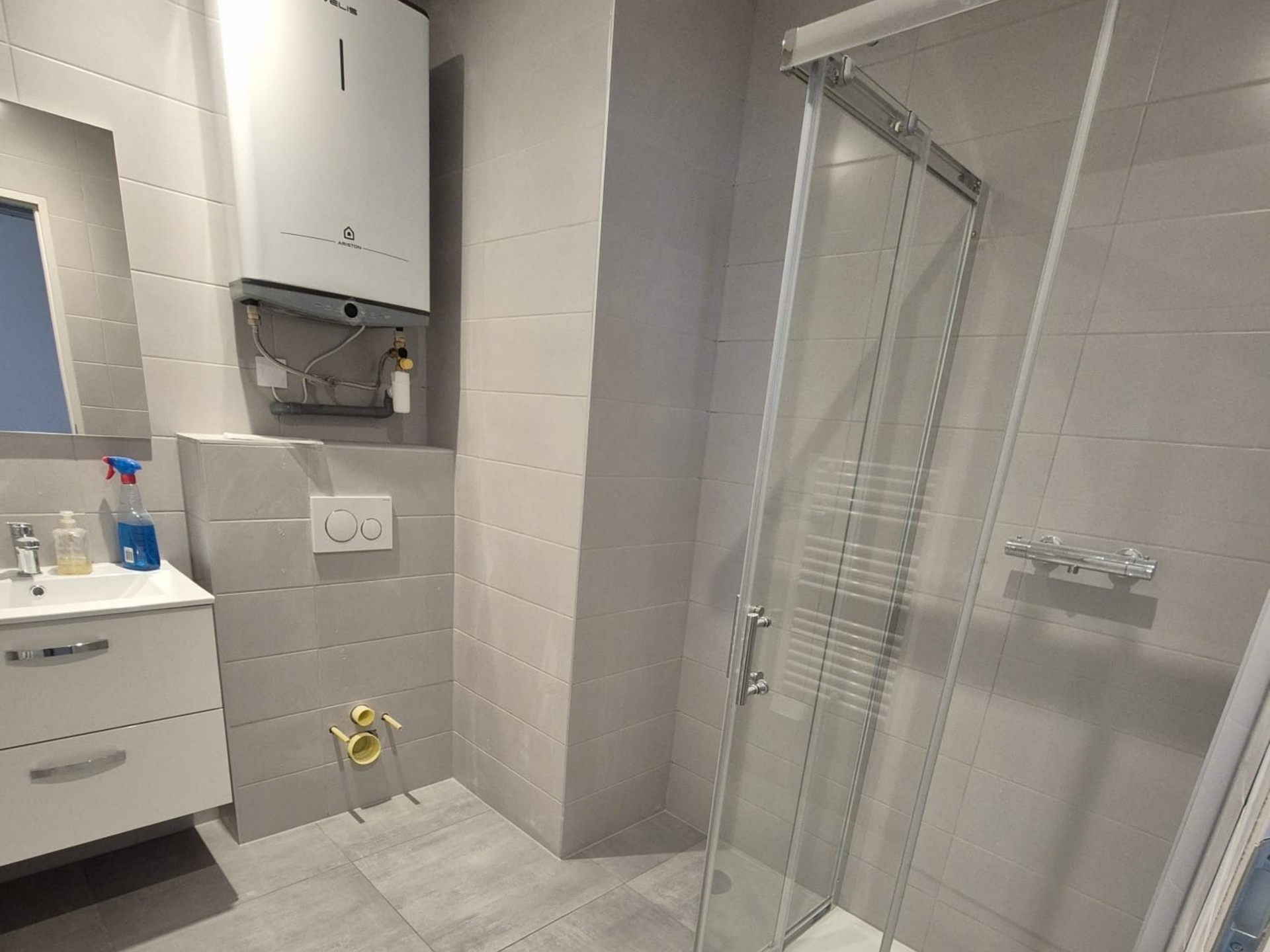 Salle de bains avec murs carrelés gris, douche, meuble lavabo et chauffe-eau.