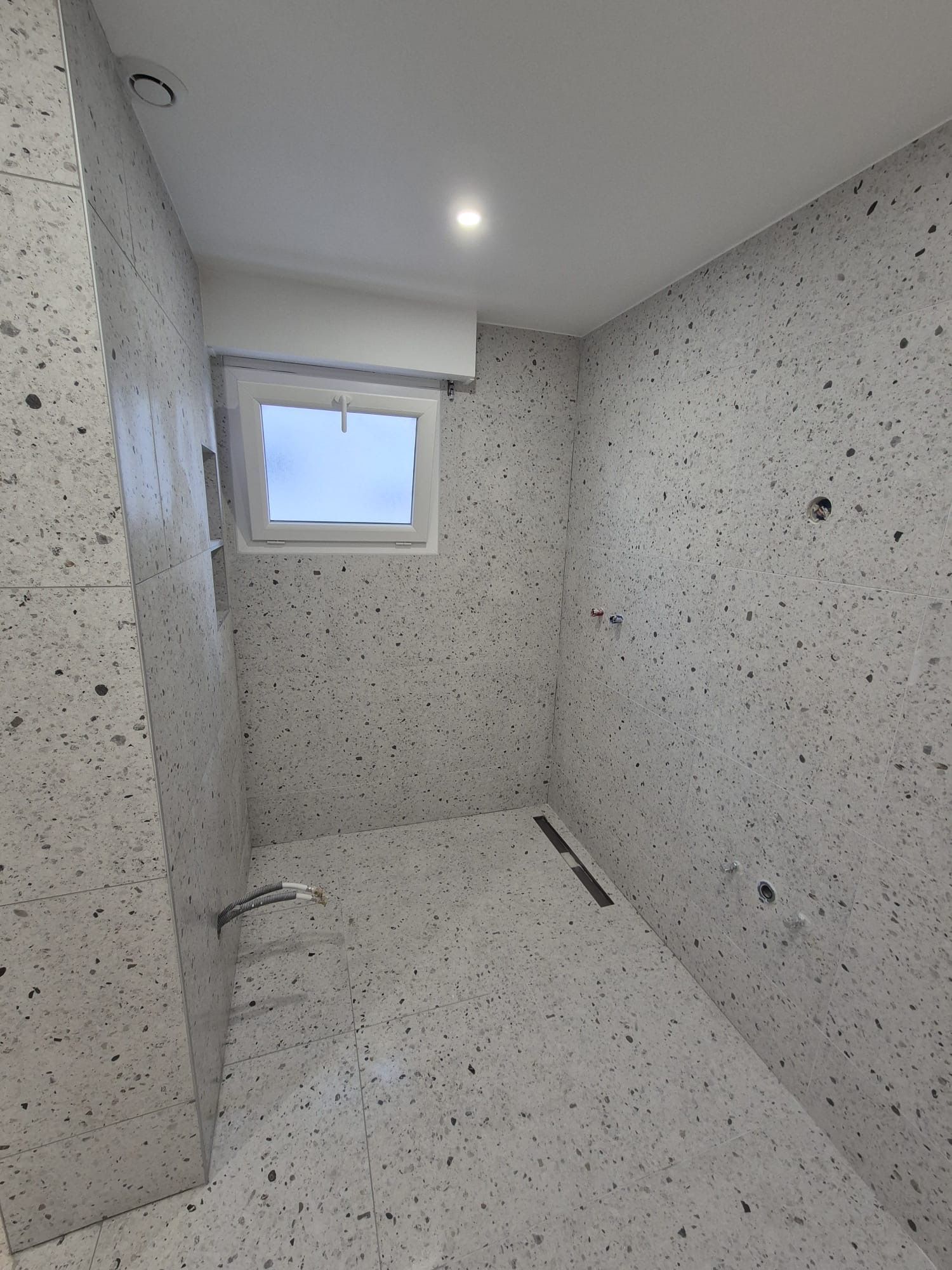 Petite salle de bains aux murs et au sol gris mouchetés, petite fenêtre et caniveaux linéaires.