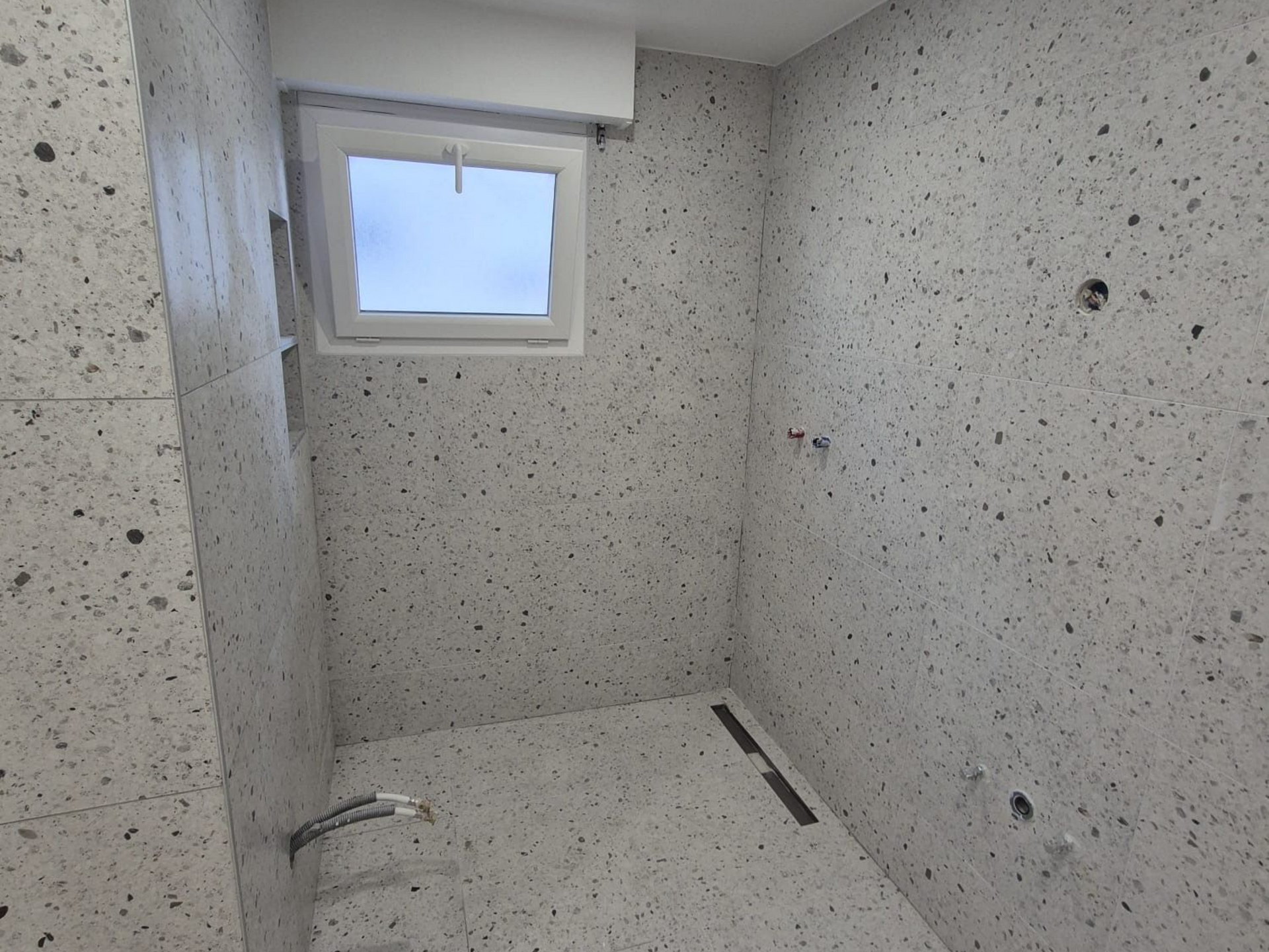 Petite salle de bains aux murs et au sol gris clair mouchetés, petite fenêtre et bonde.