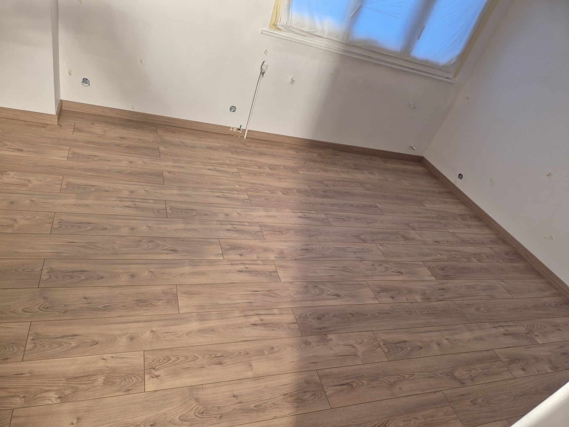 Pièce avec parquet et murs blancs, avec une fenêtre et des moulures en haut à droite.