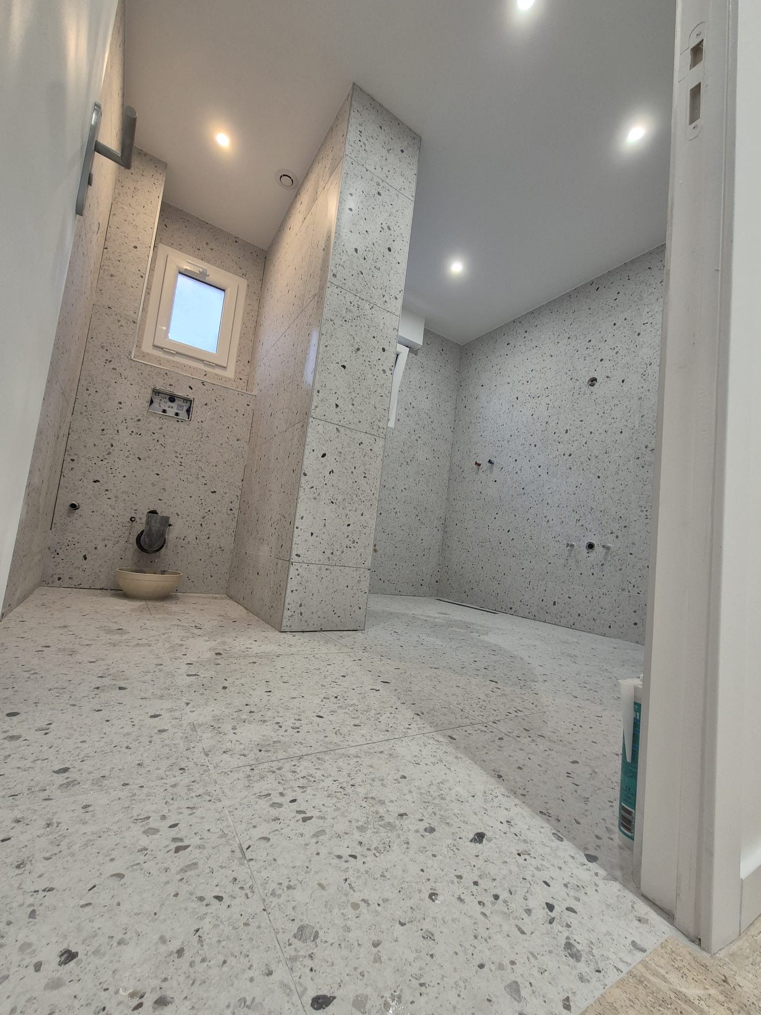 Salle de bains vide avec carrelage gris moucheté, fenêtre et éclairage au plafond.