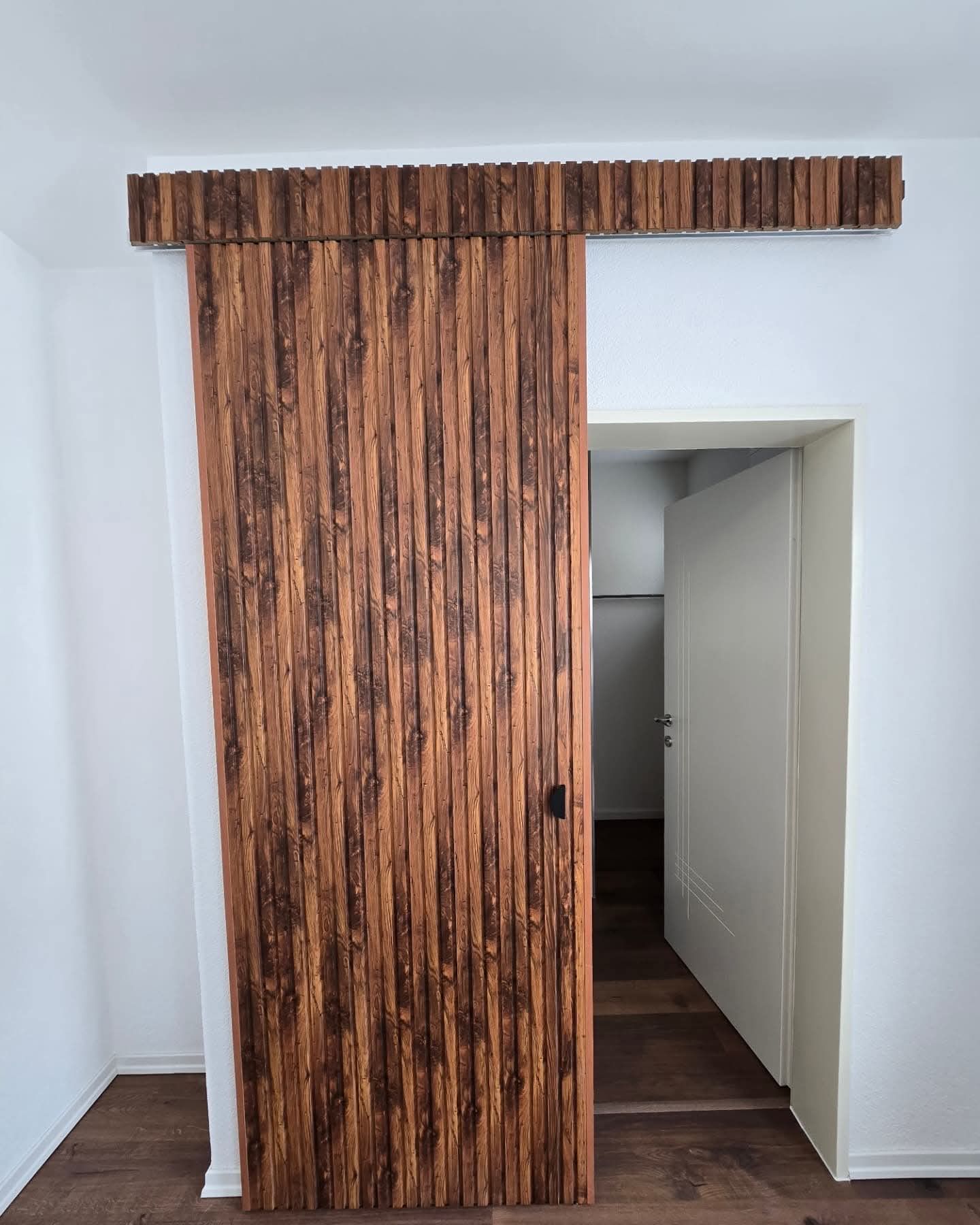 Porte coulissante en bois, entrouverte. Lattes verticales brunes au-dessus du rail. Murs blancs.