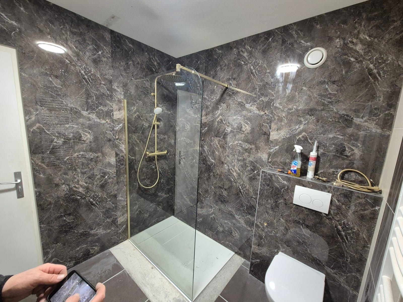 Cabine de douche avec carrelage imitation marbre foncé, robinetterie dorée, paroi de douche en verre et toilettes blanches.