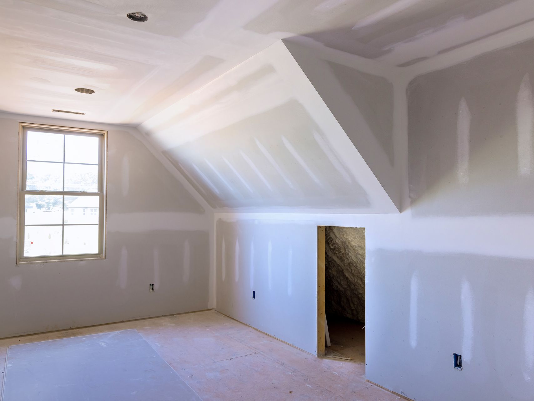 Pièce inachevée avec murs et plafond en plaques de plâtre ; fenêtre à gauche, porte à droite.