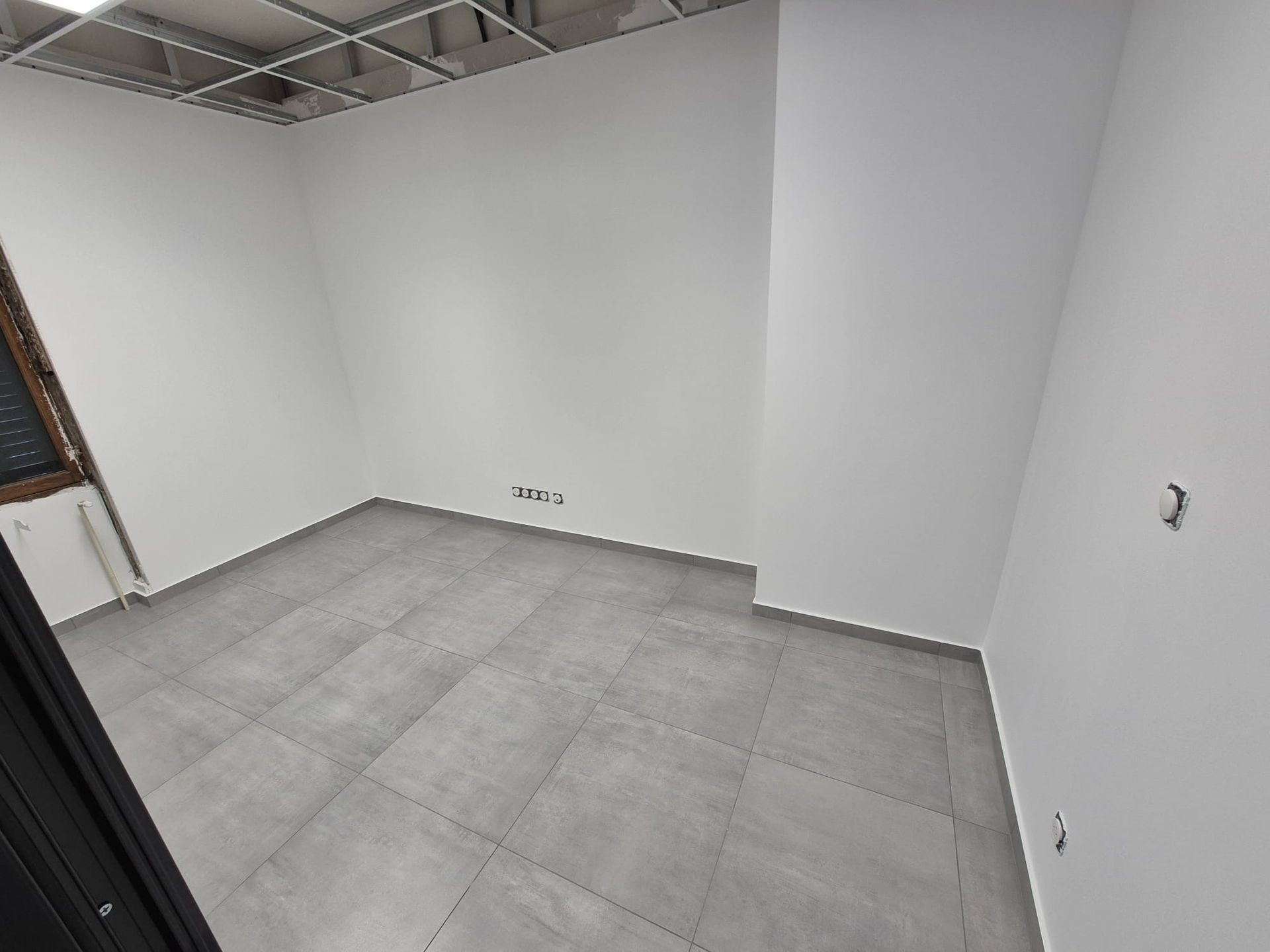 Pièce vide avec sol carrelé gris et murs blancs ; une fenêtre sur la gauche.