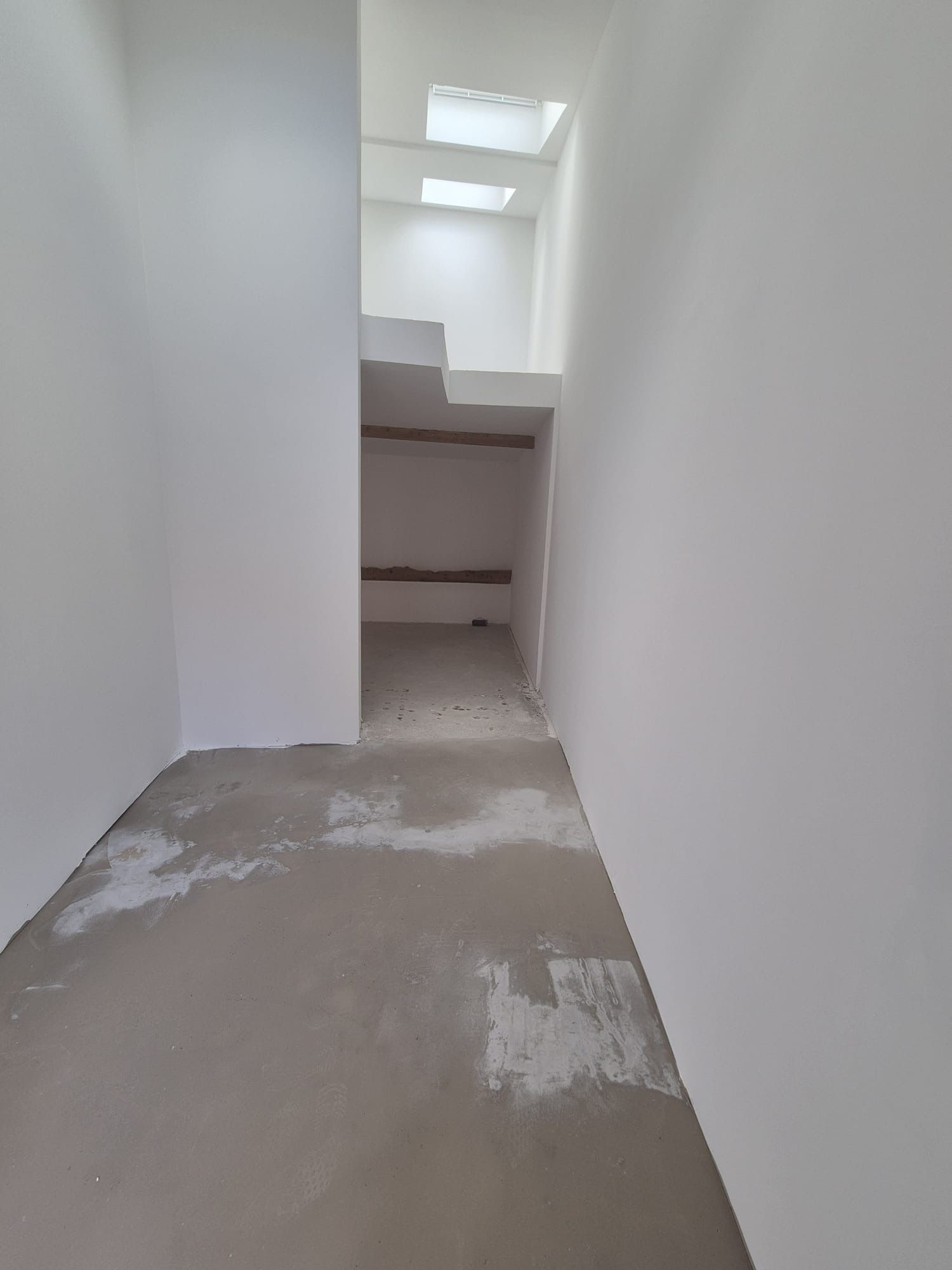 Couloir étroit et inachevé, blanc, avec sol en béton, menant à une petite ouverture et à un escalier.
