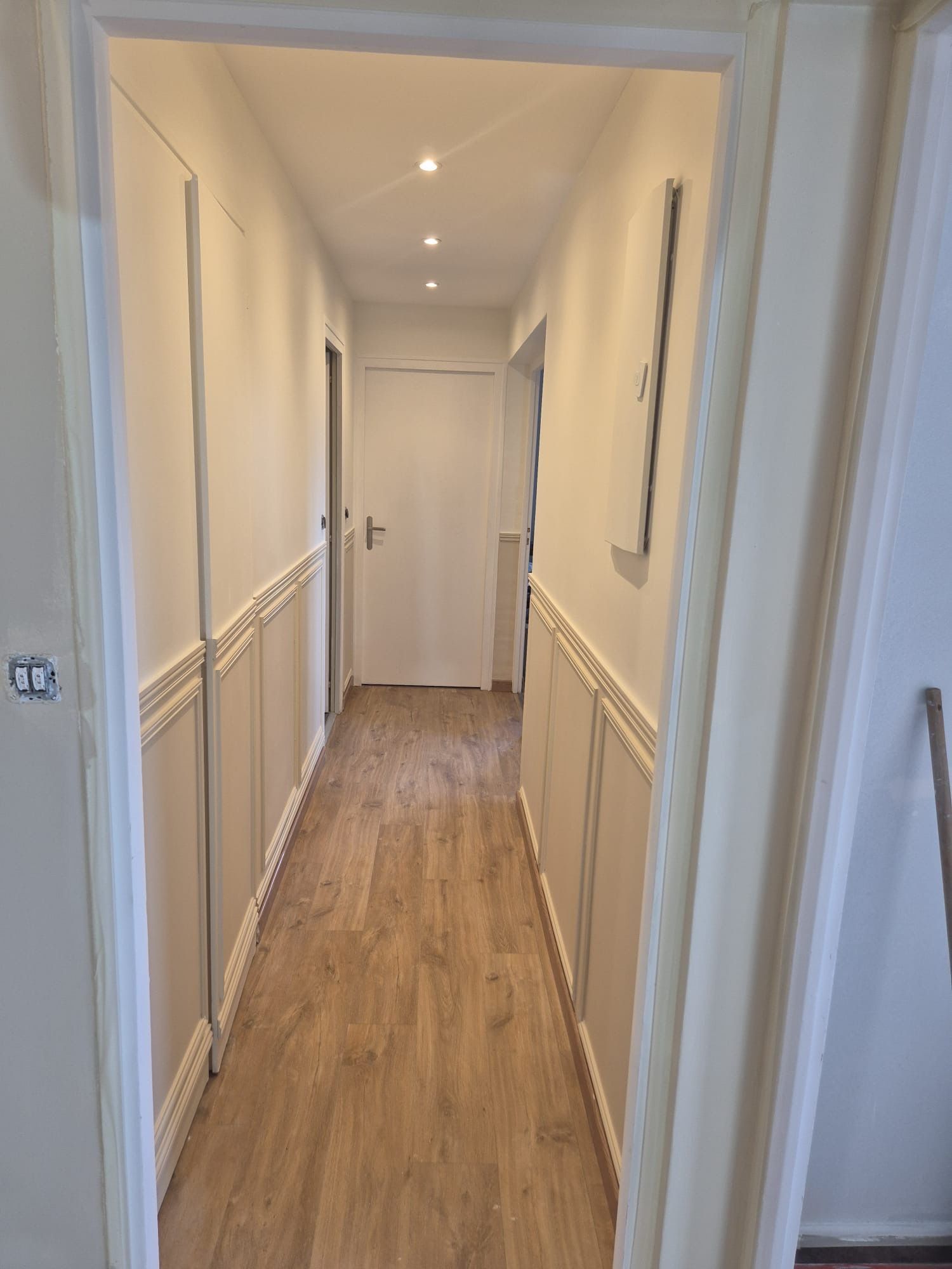 Long couloir avec parquet, murs et lambris blancs, éclairage encastré et une porte fermée au fond.