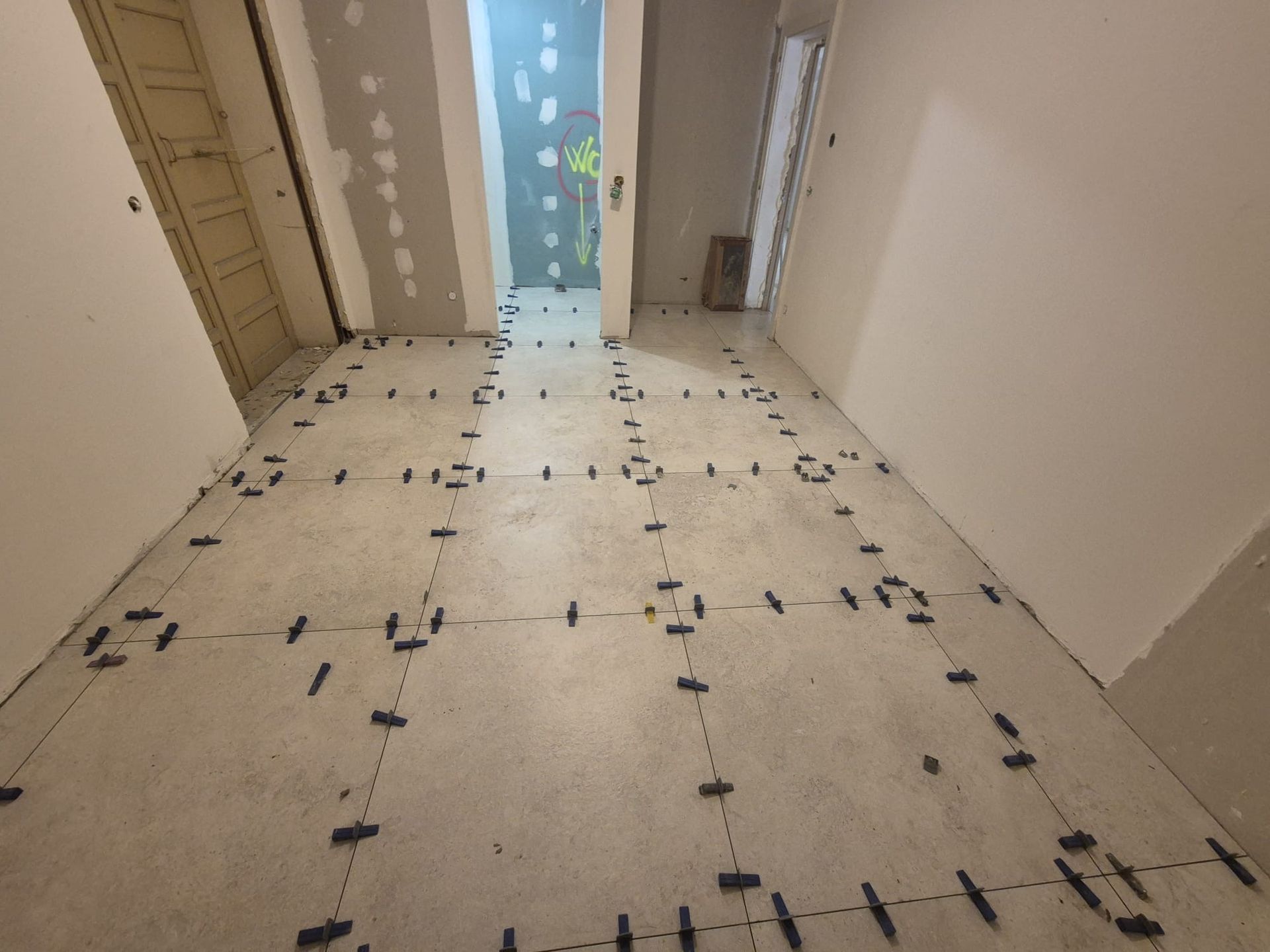 Pièce en construction, sol en béton avec clips de nivellement noirs installés, porte en arrière-plan.