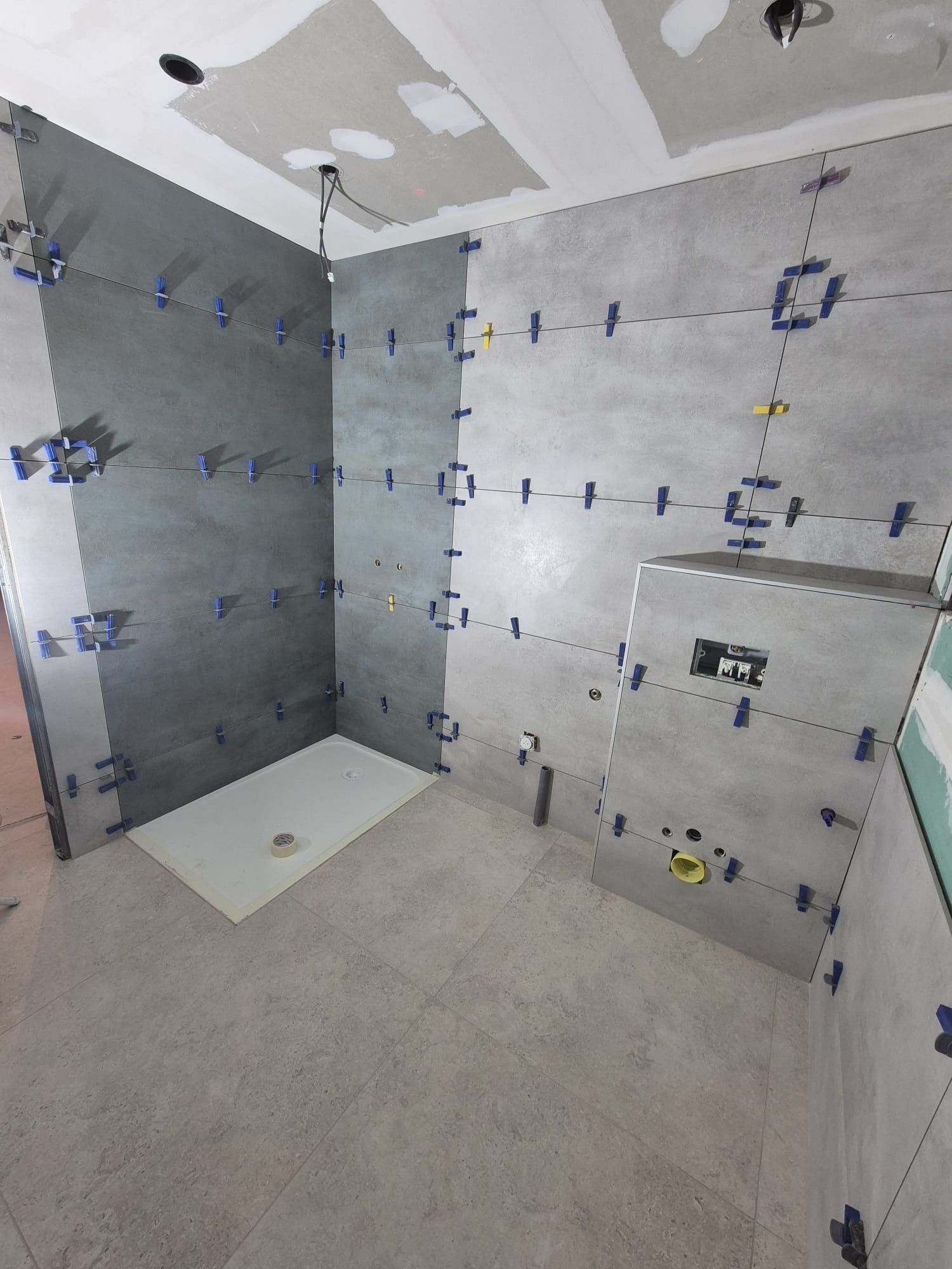 Salle de bains en construction ; carrelage gris aux murs et au sol, receveur de douche installé, clips de nivellement visibles.