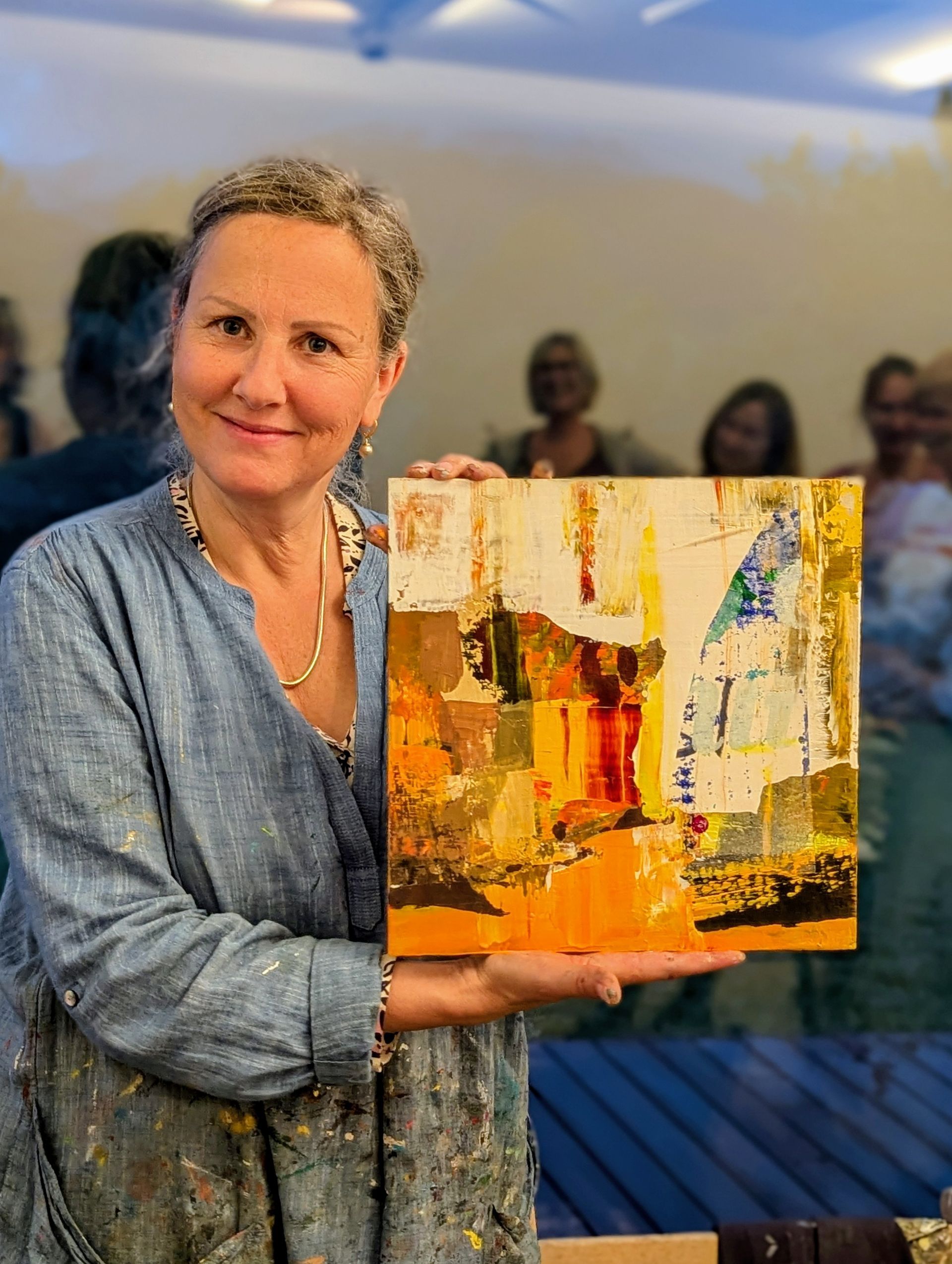 Insa Hoffman hält ein abstraktes Gemälde in Gold-, Orange- und Weißtönen; lächelt in die Kamera.