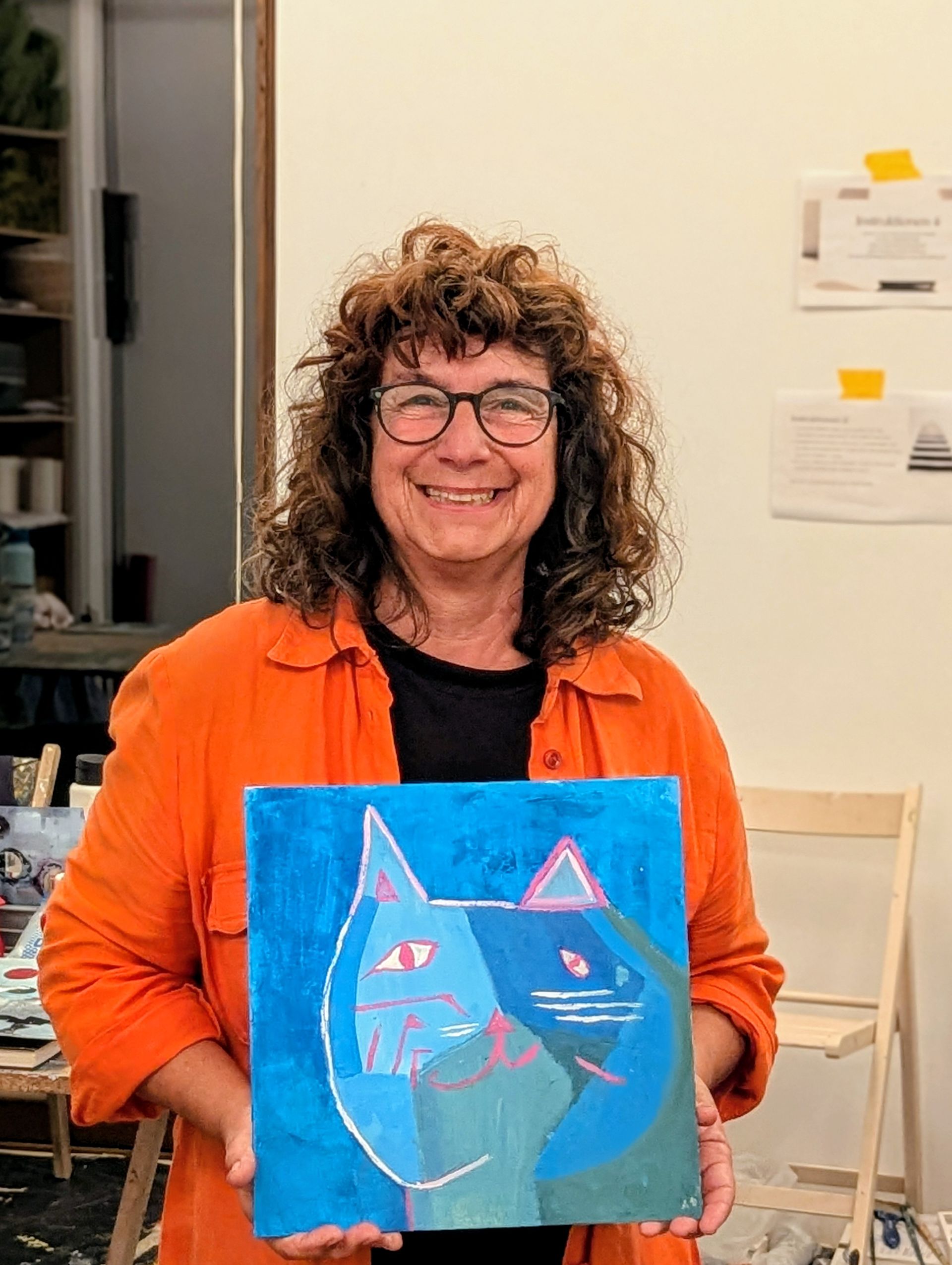 Jutta Schneider mit einem blau-türkisfarbenen Katzenbild in der Hand, die ein orangefarbenes Hemd trägt, lächelt.