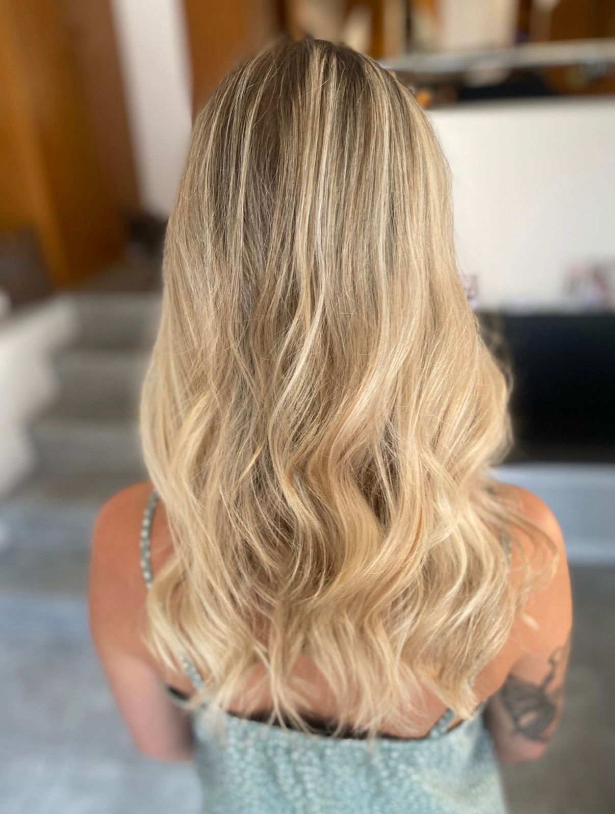 Frau mit langen, welligen blonden Haaren, Rückansicht. Sie trägt ein hellblaues Oberteil, drinnen.