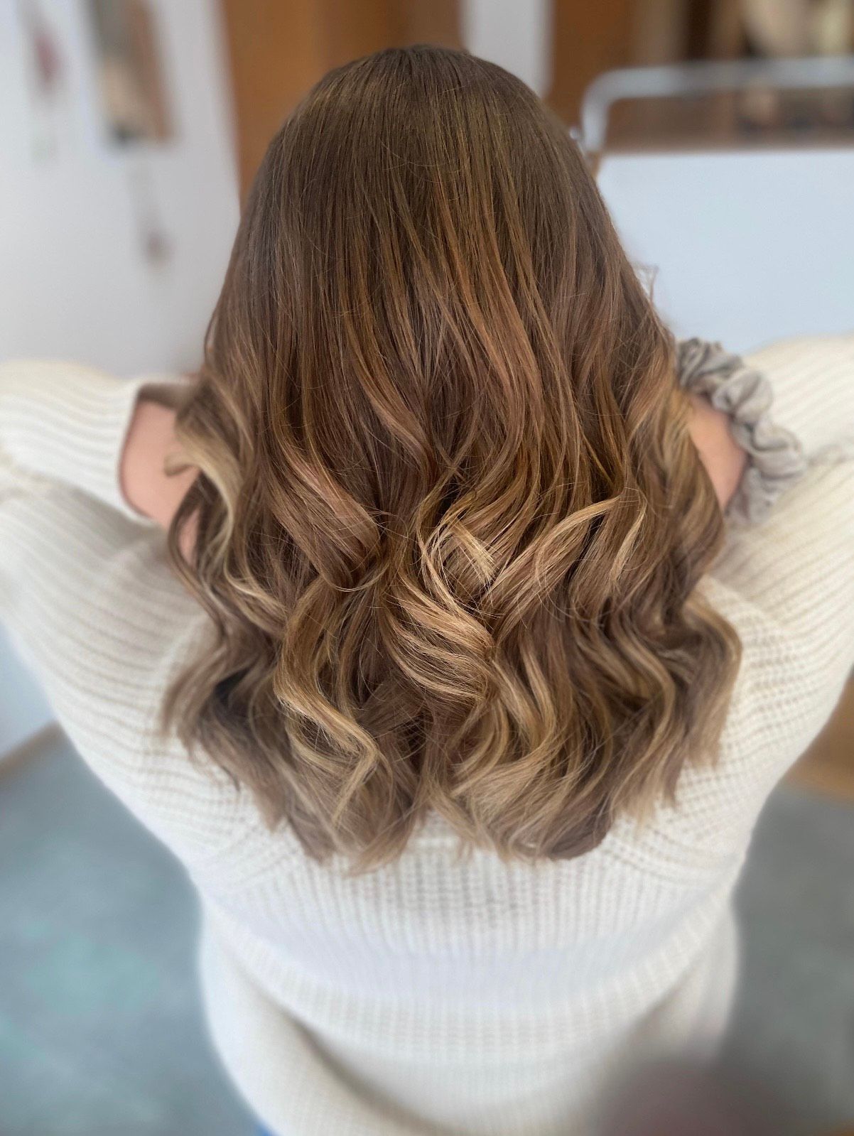 Frau mit welligem, gesträhntem braunem Haar, trägt einen weissen Pullover und hat die Hände hinter dem Kopf.