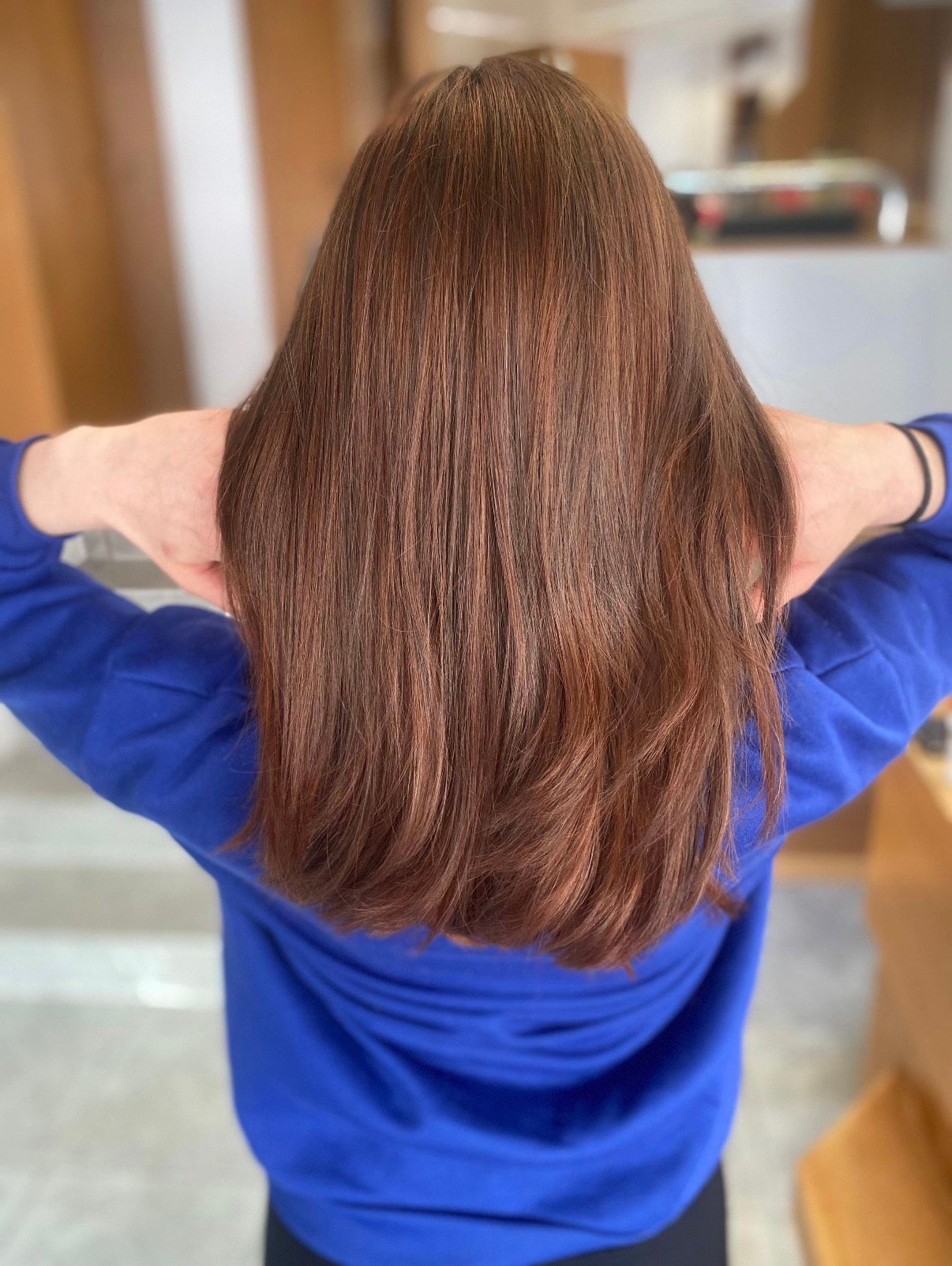 Frau mit langem, rotbraunem Haar, Hände hinter dem Kopf, trägt blauen Pullover.