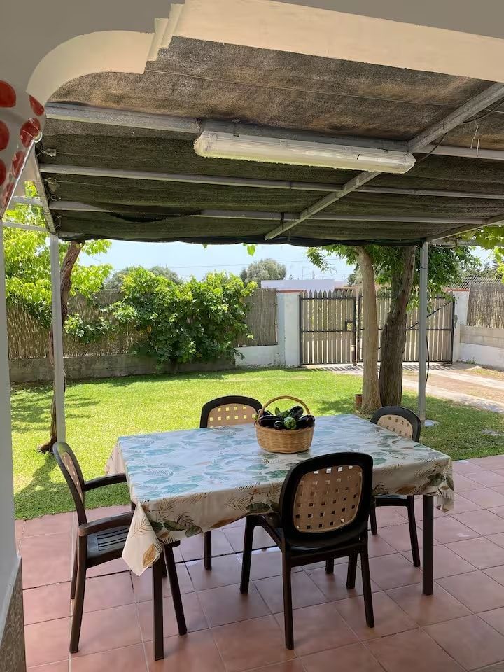 Mesa de comedor al aire libre bajo un dosel en un jardín, cuatro sillas, cesta, césped y valla.