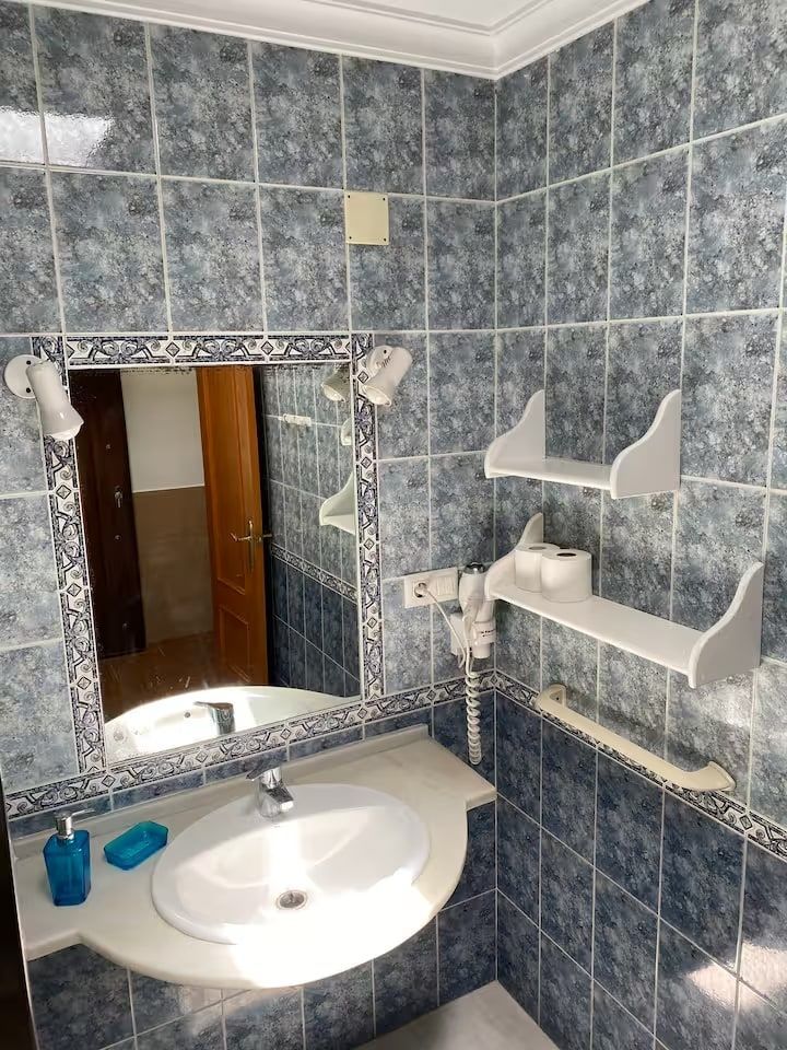 Baño con paredes de azulejos azules, lavabo con espejo, estanterías y secador de pelo.
