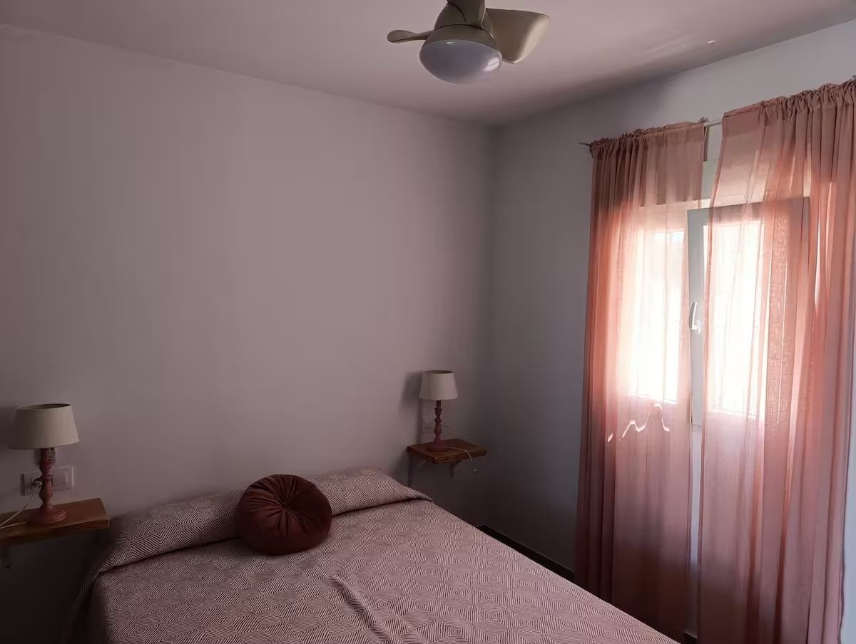 Dormitorio con cama de matrimonio, mesitas de noche pequeñas, lámparas y cortinas rosas sobre una ventana.