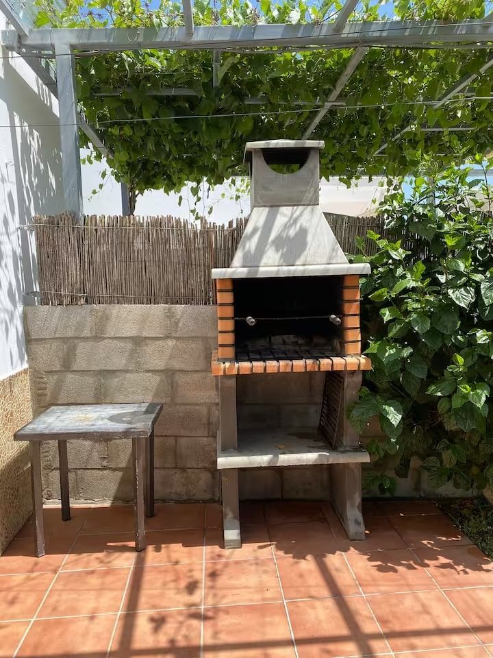 Parrilla exterior de ladrillo y hormigón con una pequeña mesa y una pérgola cubierta de enredaderas.