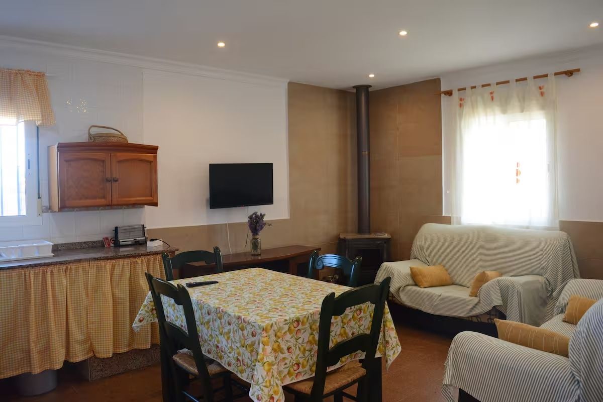 Acogedor espacio de estar con mesa de comedor, sofás, cocina y TV, luz cálida, estufa de leña.