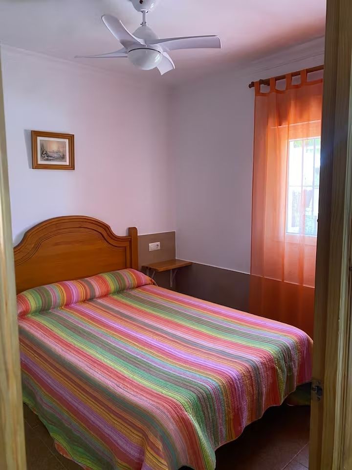 Dormitorio con una cama cubierta con una manta de rayas de colores, una cortina naranja y un ventilador de techo.