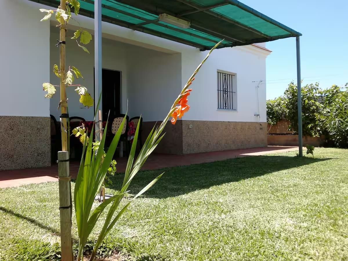 Casa blanca con toldo verde, flores naranjas en el patio, cielo azul.