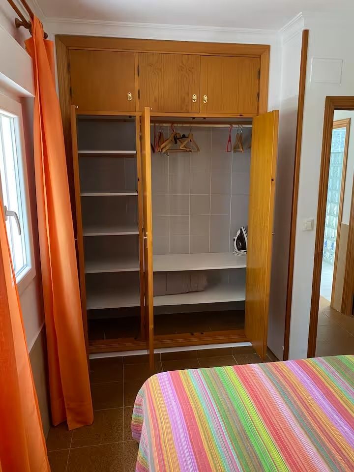 Armario de dormitorio con puertas abiertas, estantes y perchas. Cortinas naranjas y colcha colorida.